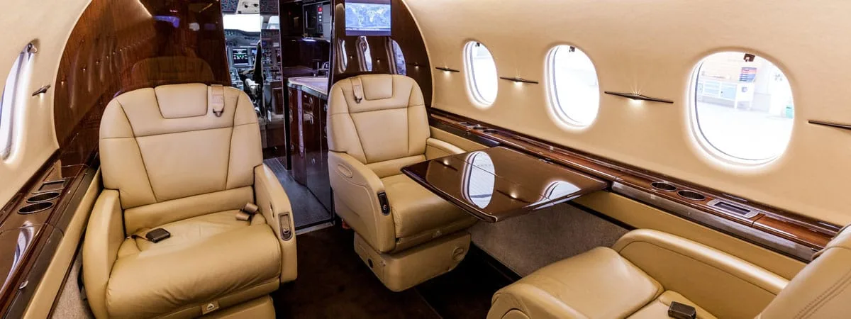 A wide shot of an aircraft's luxurious cabin interior featuring tan leather seats, a polished wood table, and a view towards the cockpit.
