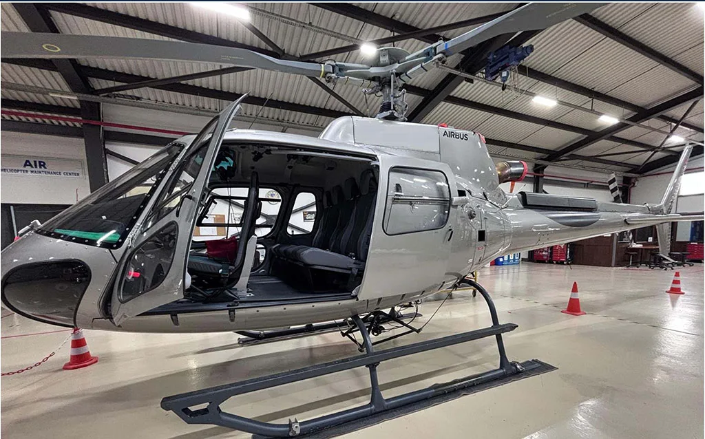 A silver Airbus H125 helicopter with its doors open, parked inside a hangar.