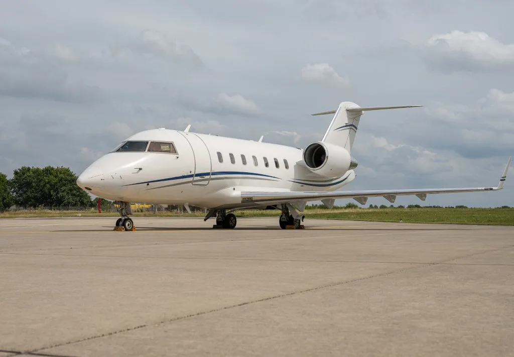 A white Bombardier Challenger business jet with blue and grey stripes on the tarmac under a cloudy sky.