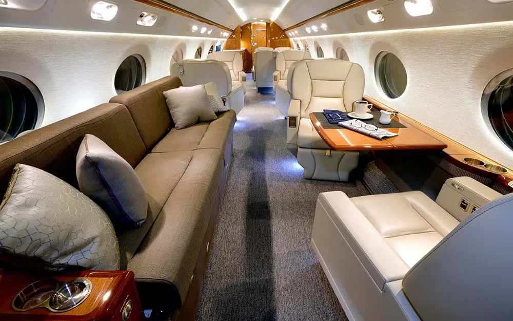 A luxurious aircraft cabin interior featuring a long brown sofa, beige leather seats, and a table set with coffee and a newspaper.