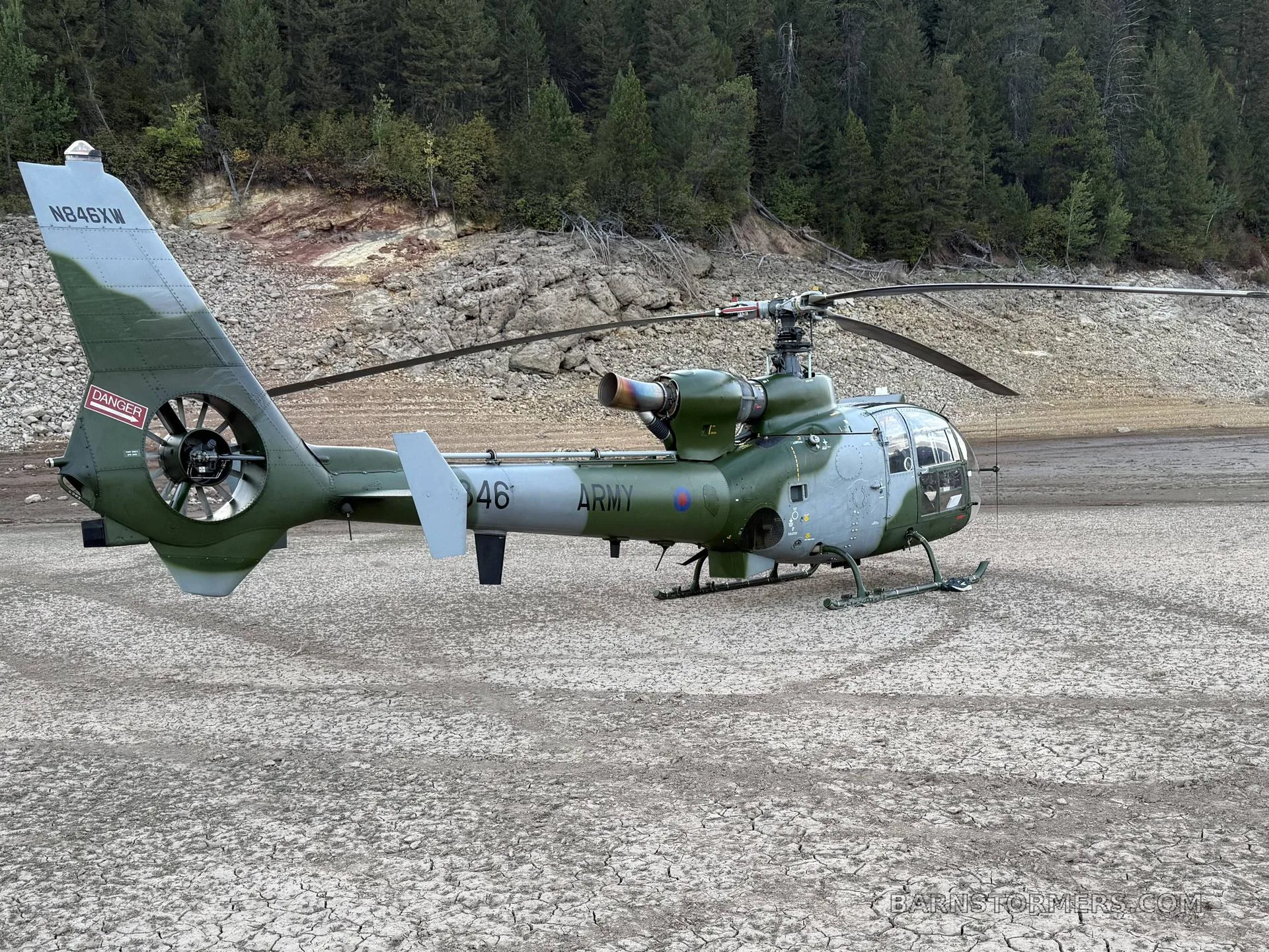 A green and grey military Westland Gazelle helicopter is seen from the rear on dry, cracked earth with a forest in the background.