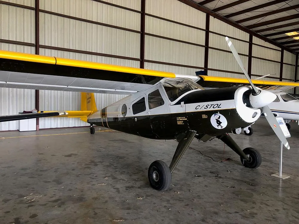 A dark green and white utility aircraft with yellow wings and tail, featuring a 'C/STOL' marking and a cartoon decal on the nose, parked inside a hangar.