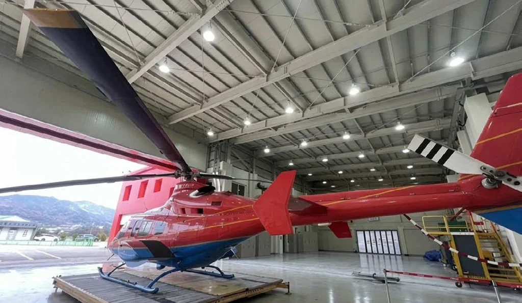 A red and blue helicopter is seen from the rear-side, with its tail rotor visible, partially in a hangar.