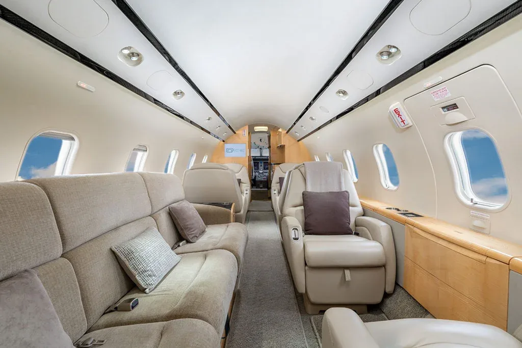 The interior cabin of a private jet featuring a beige sofa, individual seats, and light wood paneling.