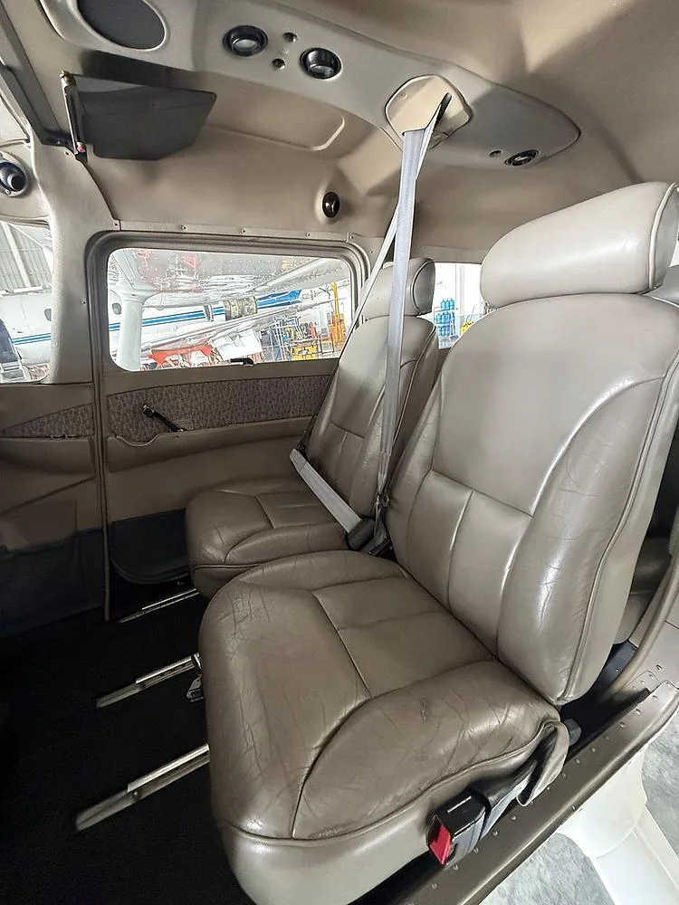The tan leather rear passenger seats of a small aircraft cabin are visible, with a view of other aircraft outside the window.