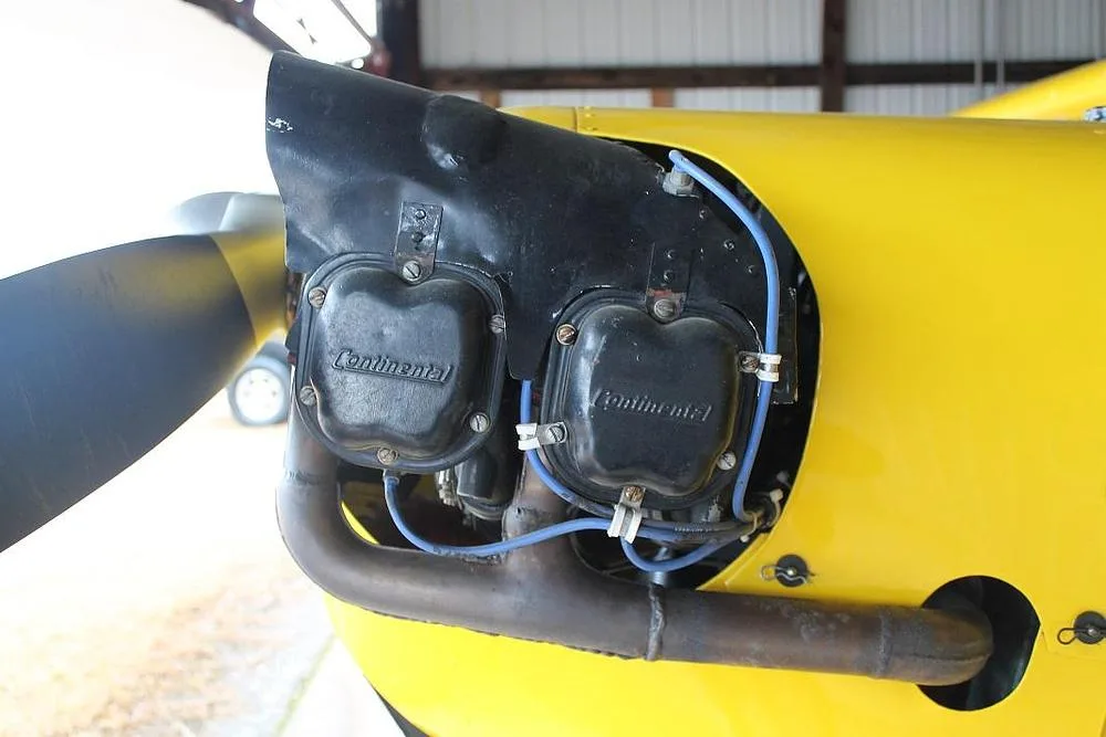 A close-up view of the Continental engine on the yellow aircraft, showing the exhaust pipe and blue ignition wires.