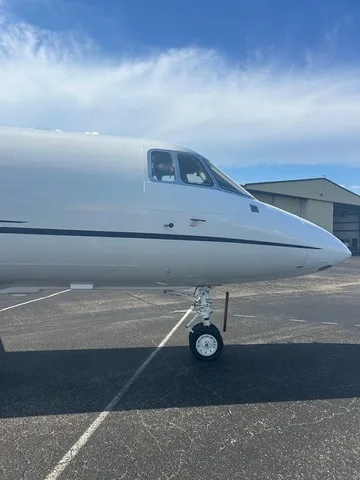 A close-up view of the front fuselage and cockpit windows of a white business jet with a dark stripe, parked on an asphalt tarmac.