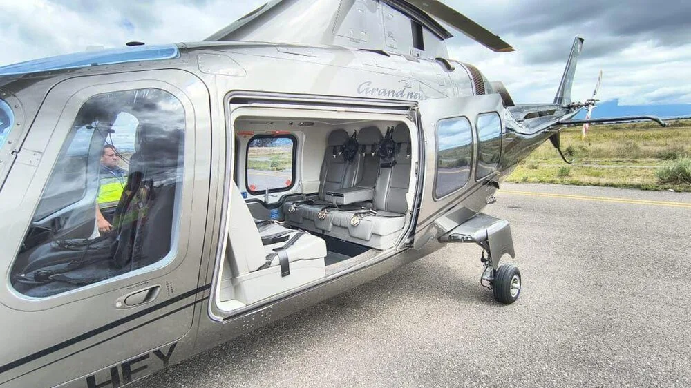 A dark grey Leonardo AW109 GrandNew helicopter is parked with its cabin door open, revealing the grey passenger seats inside and a pilot in the cockpit.