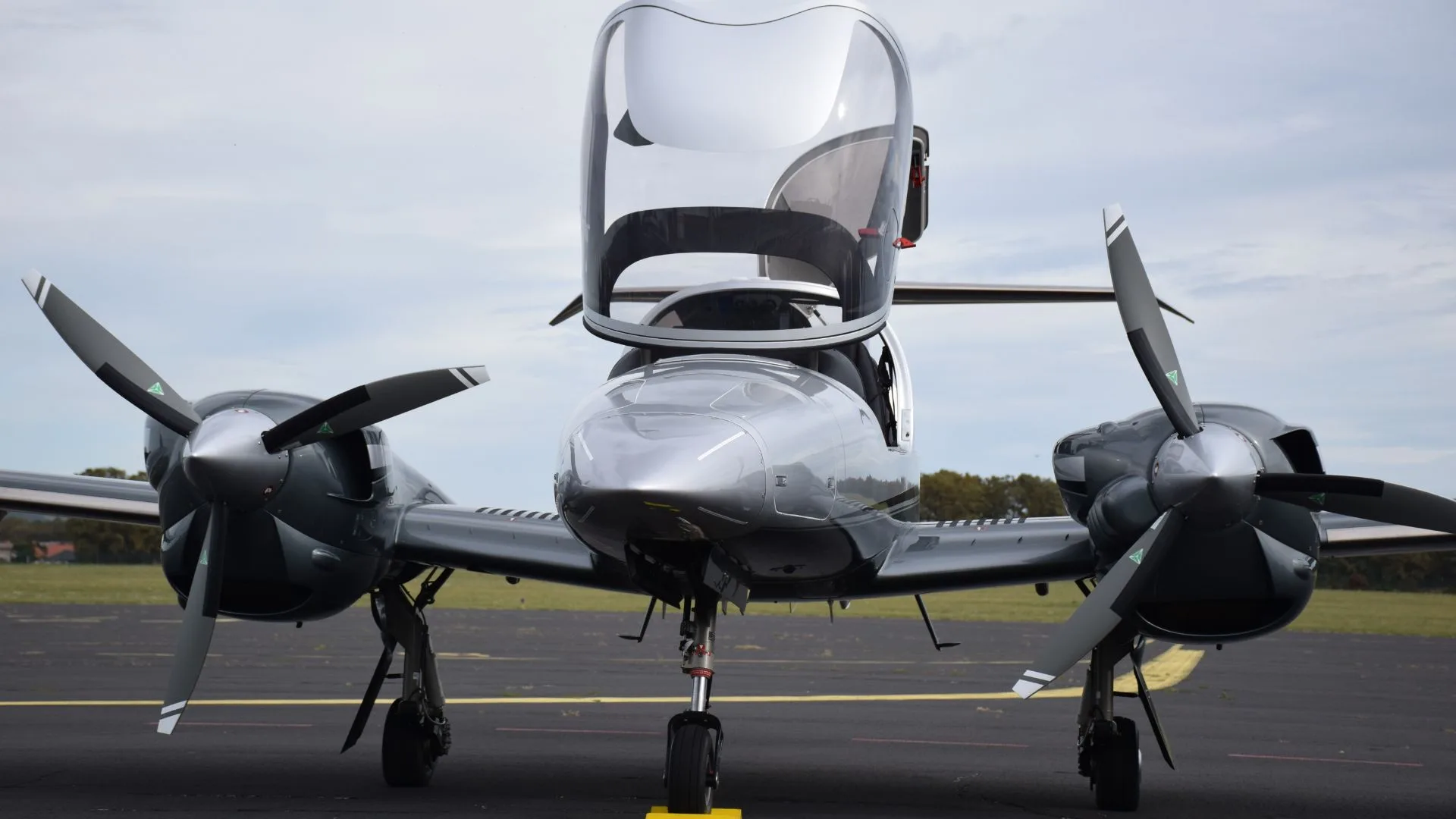 A front view of a silver Diamond DA62 twin-engine aircraft with its canopy open on an airfield.