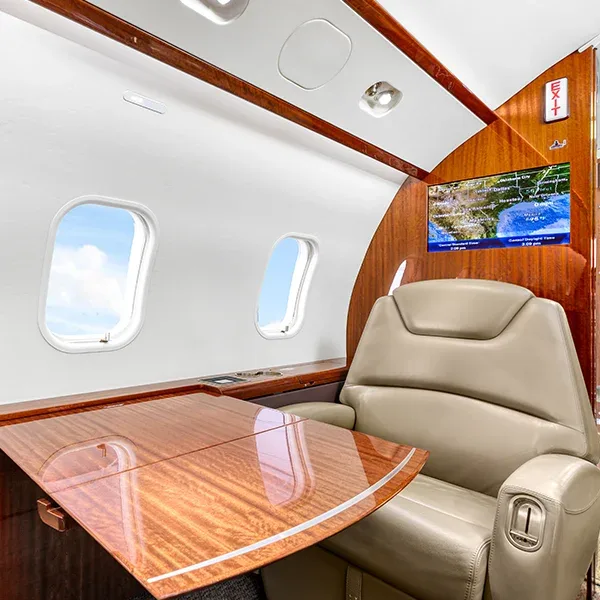 A luxurious aircraft cabin interior featuring a beige leather seat, a polished wooden table, and a monitor displaying a map.