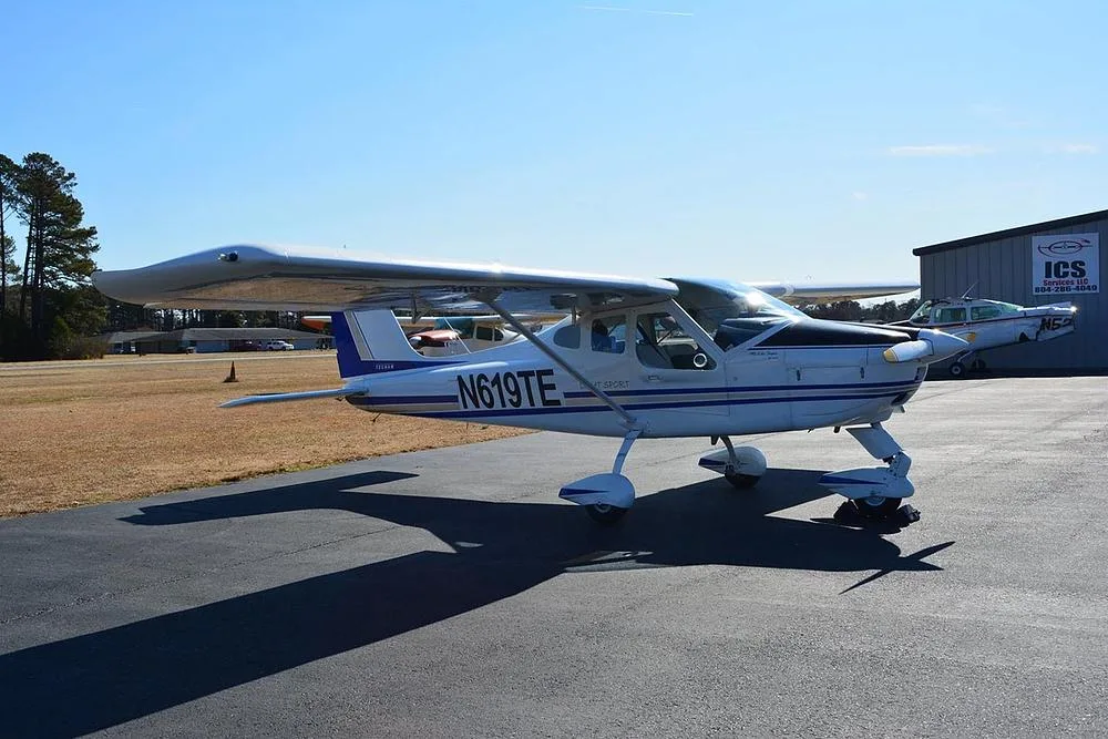 A white Tecnam Light Sport aircraft with blue and grey stripes parked on an asphalt ramp next to a hangar.