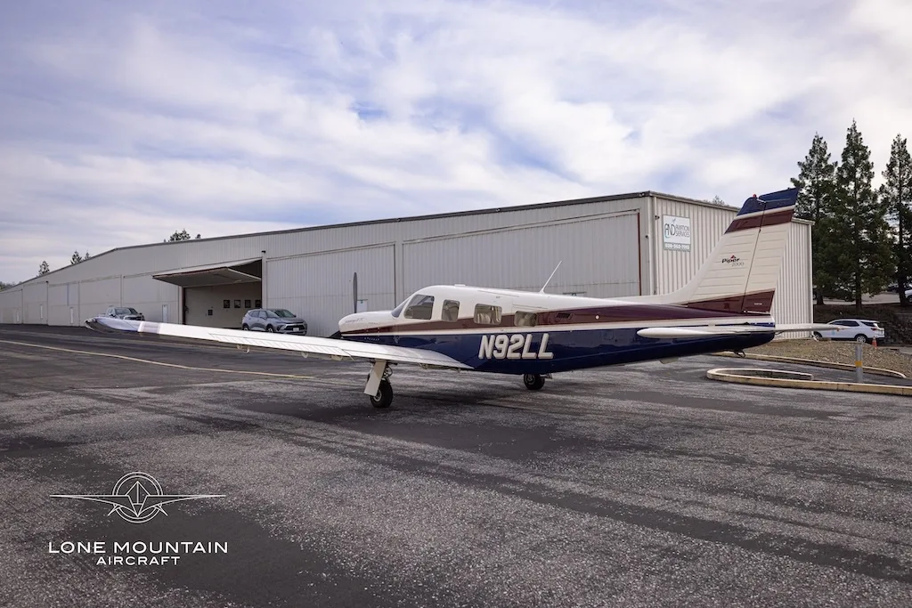 A three-quarter rear view of the Piper Saratoga II TC aircraft (N92LL) with blue and white paint scheme parked on an asphalt apron near a hangar.