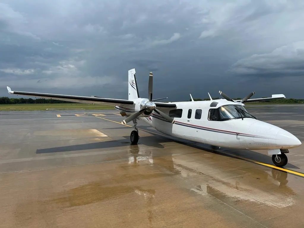 A white twin-engine turboprop aircraft with red and blue stripes is parked on a wet tarmac under a dramatic cloudy sky.