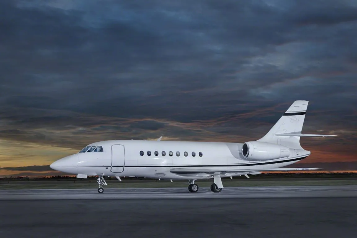 A white Dassault Falcon business jet seen from the side on a runway at sunset.