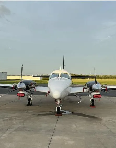 A head-on view of a white and black twin-engine propeller aircraft with red propeller covers, parked on a tarmac.