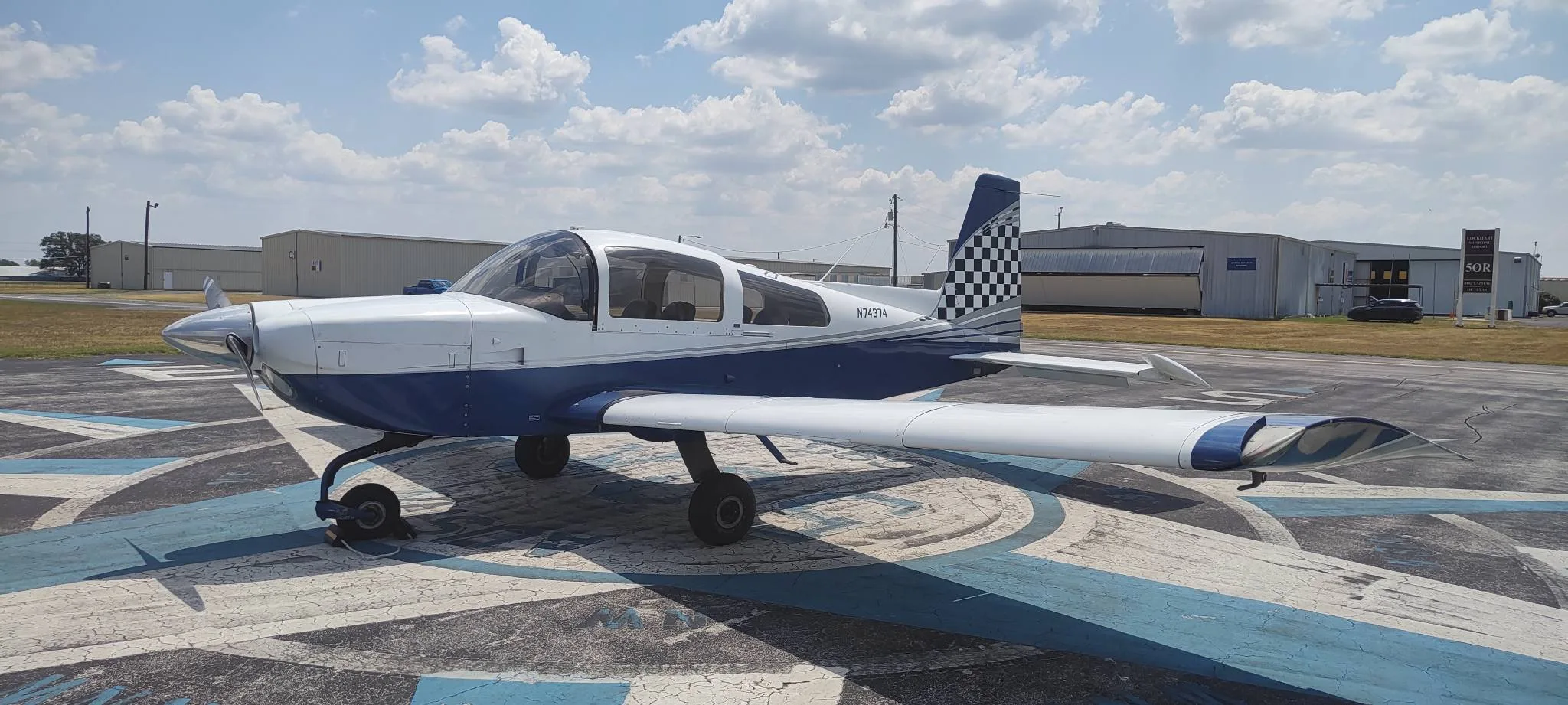A blue and white single-engine aircraft, N74374, with a checkered tail, parked on an airfield compass rose.