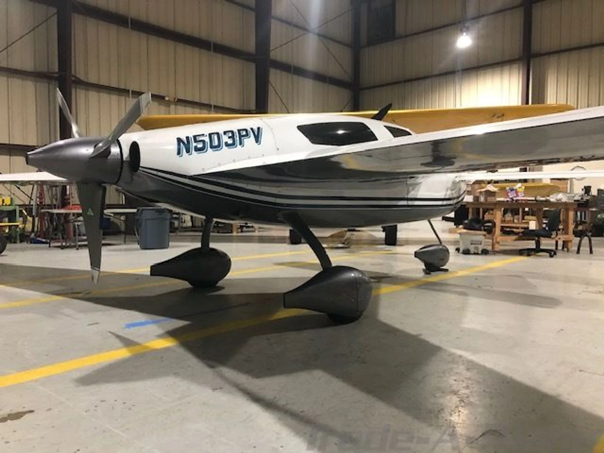 Three-quarter front view of a white and dark grey Velocity aircraft with a propeller, parked in a hangar.