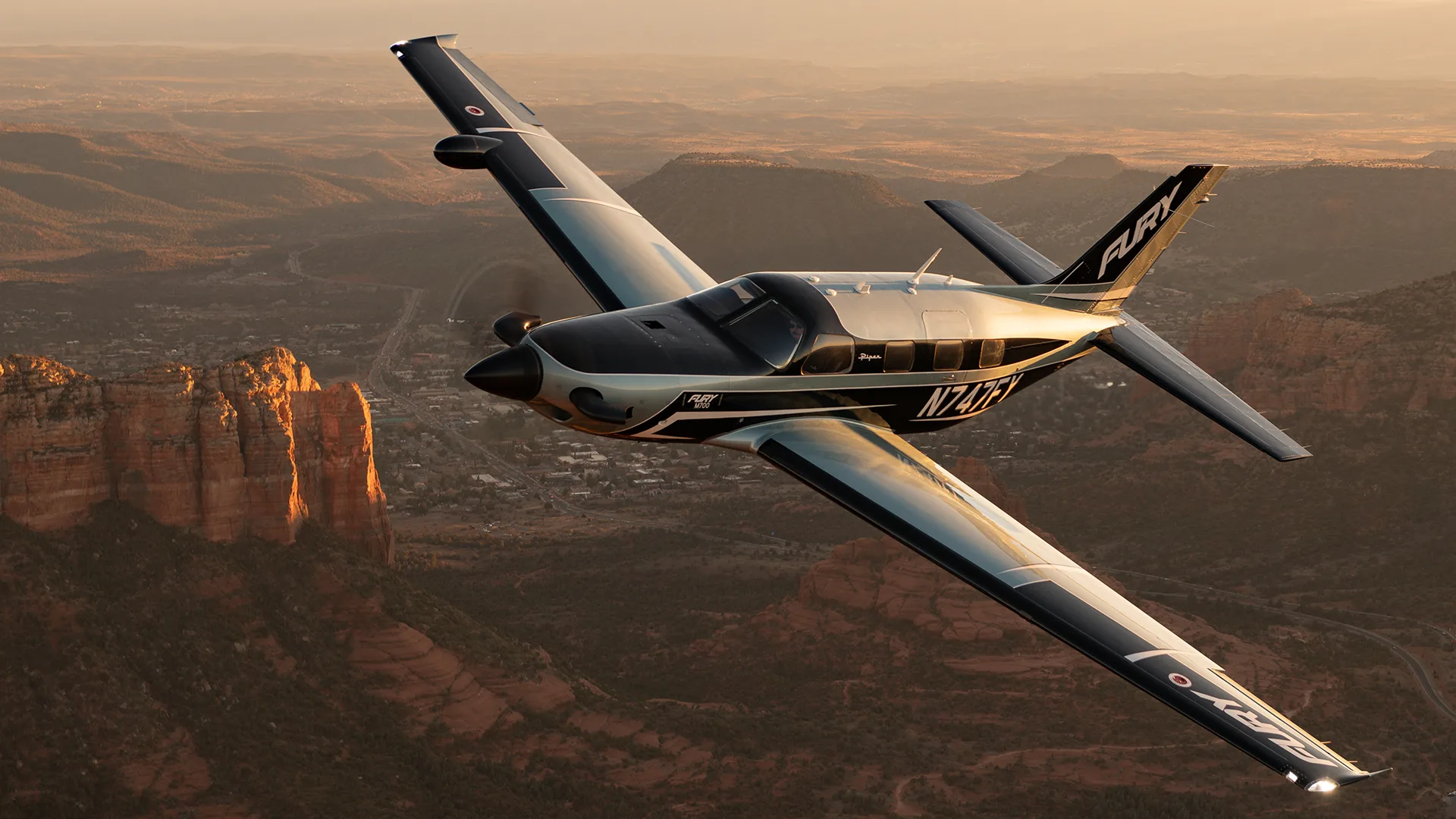 A black and silver Piper M700 Fury aircraft with tail number N747FX flying over a desert landscape with red rock formations.