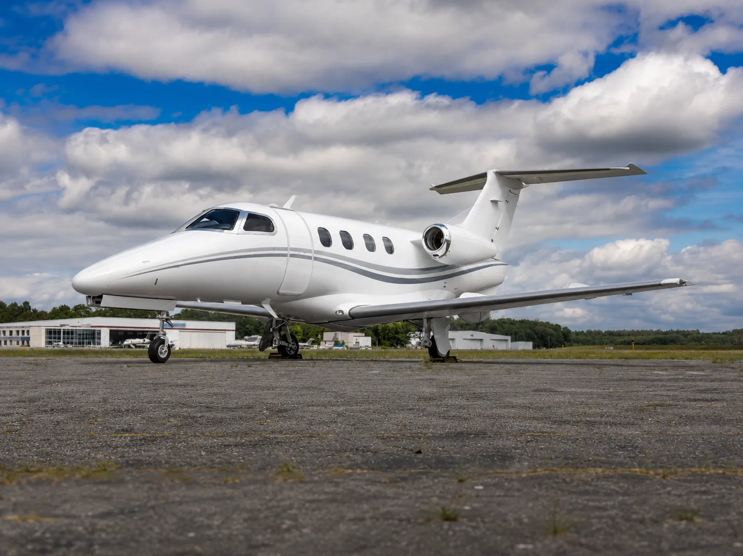 A white business jet with grey stripes is parked on an asphalt tarmac under a partly cloudy sky.
