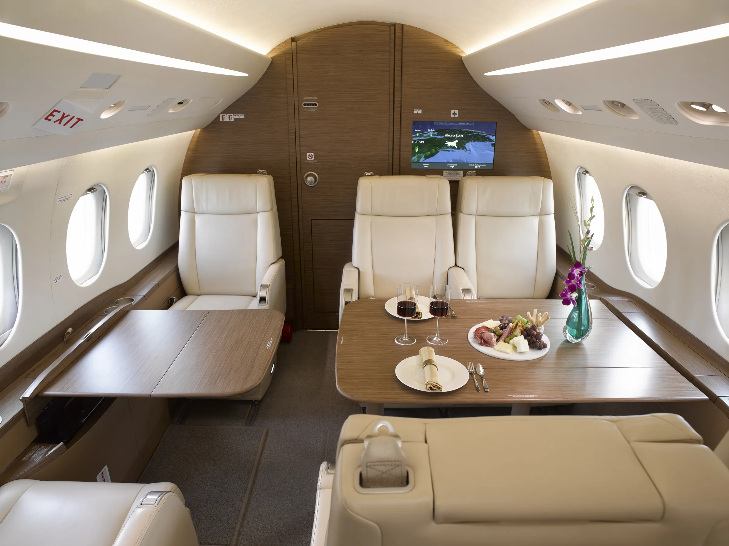 A luxurious aircraft cabin interior with cream leather seats, a wooden table set with food and drinks, and a monitor displaying a map.