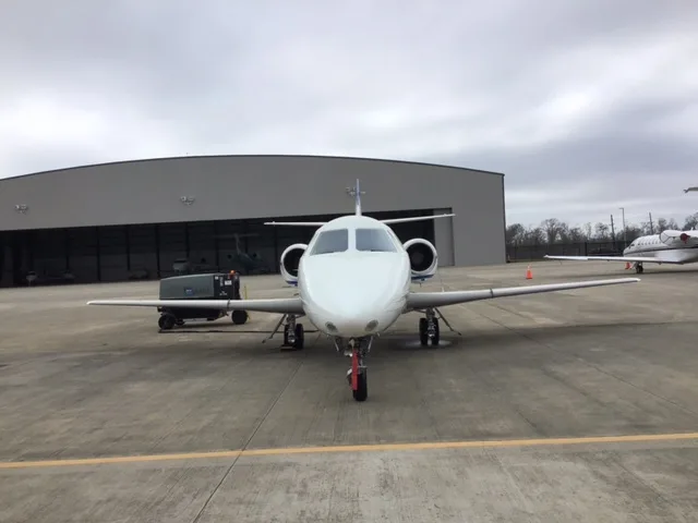 The front view of a white business jet with blue and grey stripes, parked on an airfield in front of a hangar.