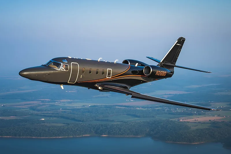 A dark gray jet with orange stripes, registration N26BD, in flight over a rural landscape.