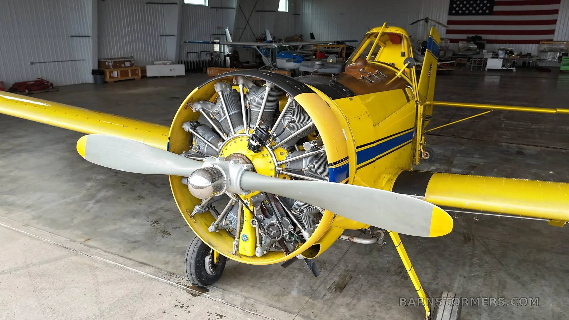 A yellow agricultural aircraft with its radial engine exposed, parked inside a hangar with an American flag in the background.