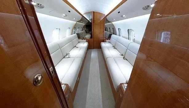 A perspective view down the aisle of a private jet cabin, flanked by two long beige leather sofas and rich wooden walls.