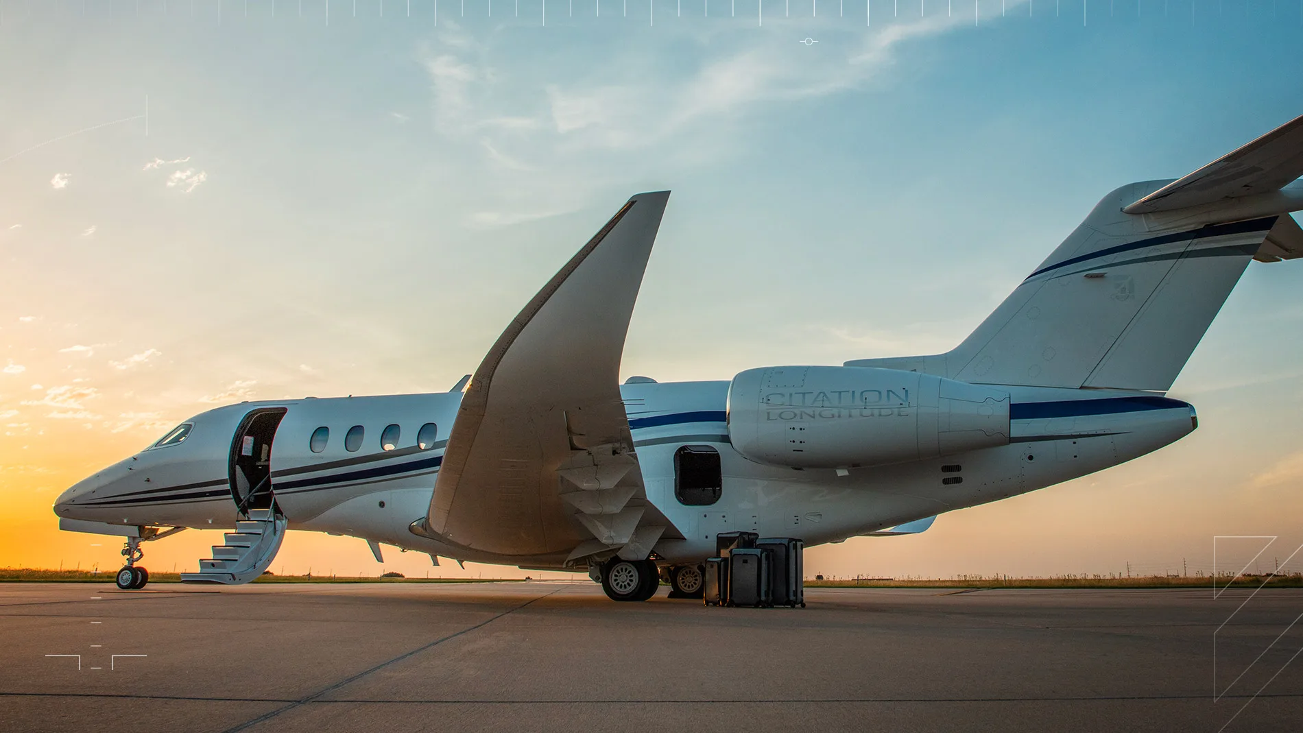 A white Cessna Citation Longitude private jet with blue stripes is parked on an airfield at sunset, with its main door open and stairs extended, and luggage on the tarmac.