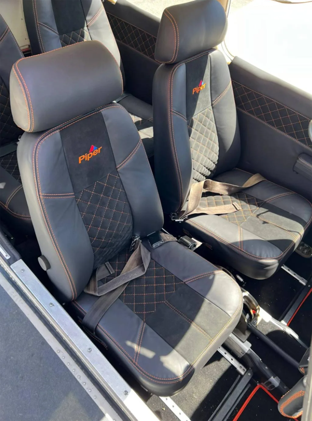 Two black leather aircraft seats with orange stitching and "Piper" logo on the headrests.