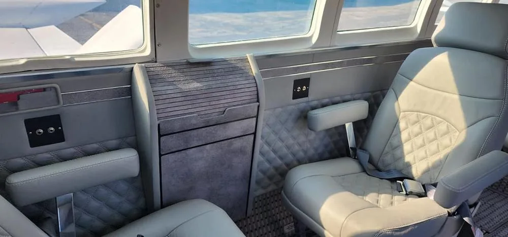 A close-up of the aircraft cabin interior showing a light gray quilted leather seat, a side table, and wood-paneled walls.