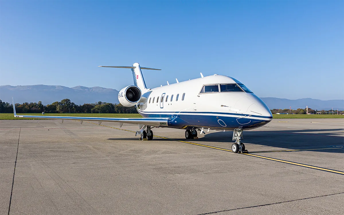 A white and blue Bombardier Challenger 604 business jet is parked on an airport tarmac with mountains in the background under a clear sky.