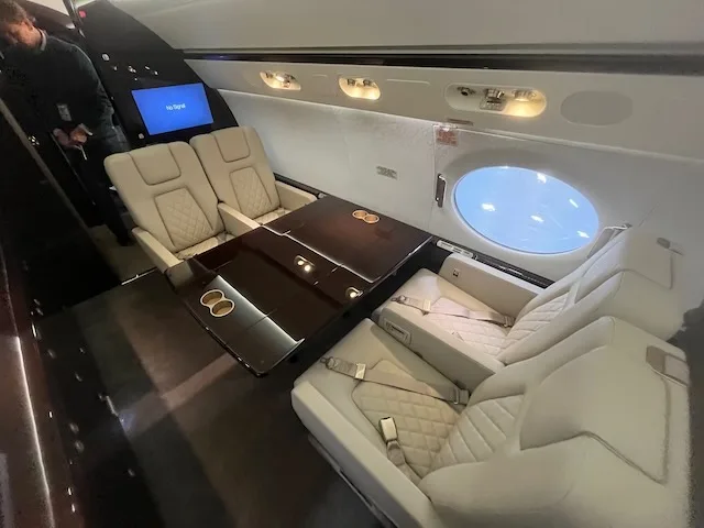 The luxurious interior of a business jet cabin with light-colored quilted seats and a dark wood table.