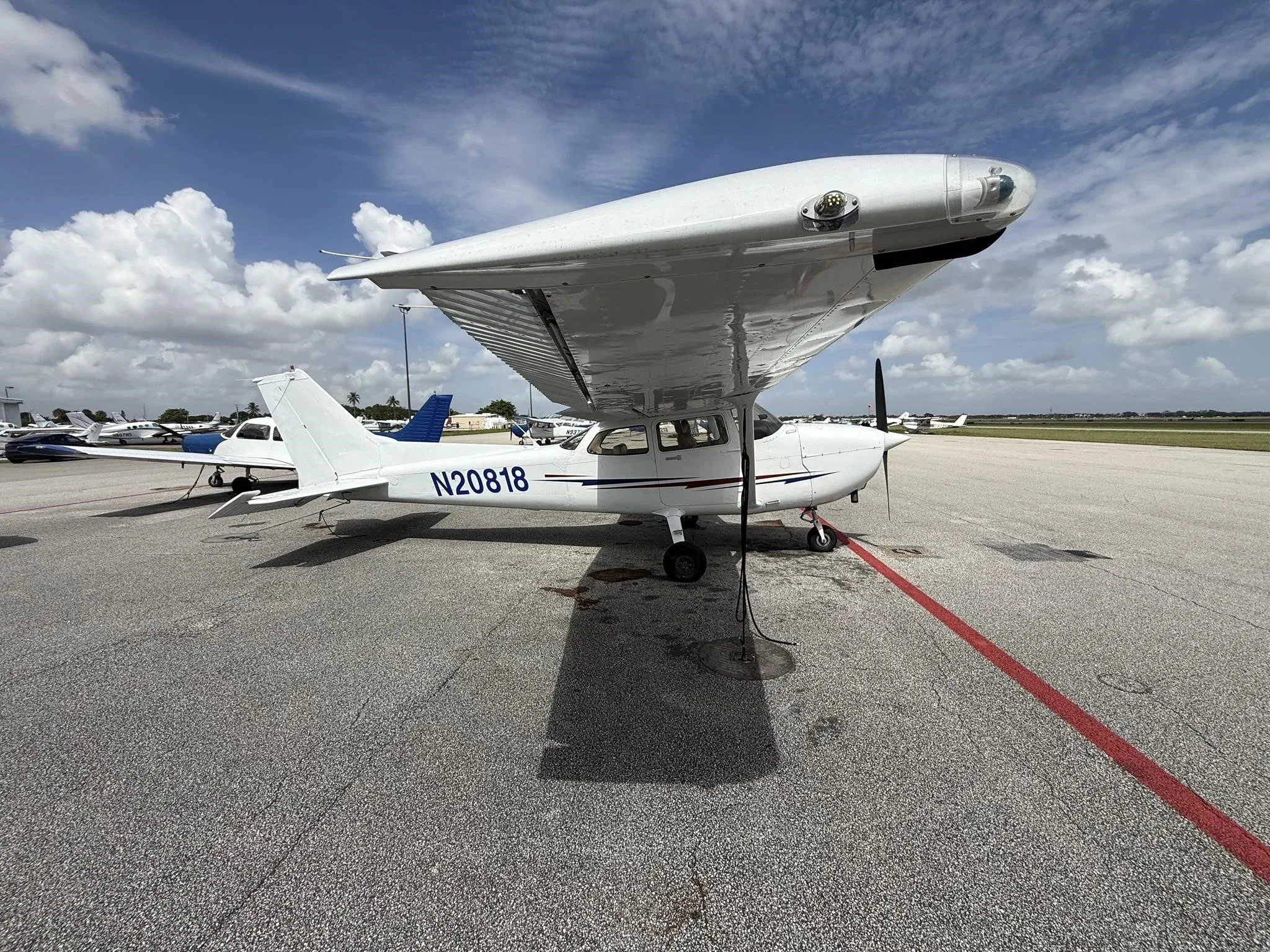 A white Cessna 172 (N20818) parked on an airport tarmac, viewed from the front-left with other aircraft in the background.