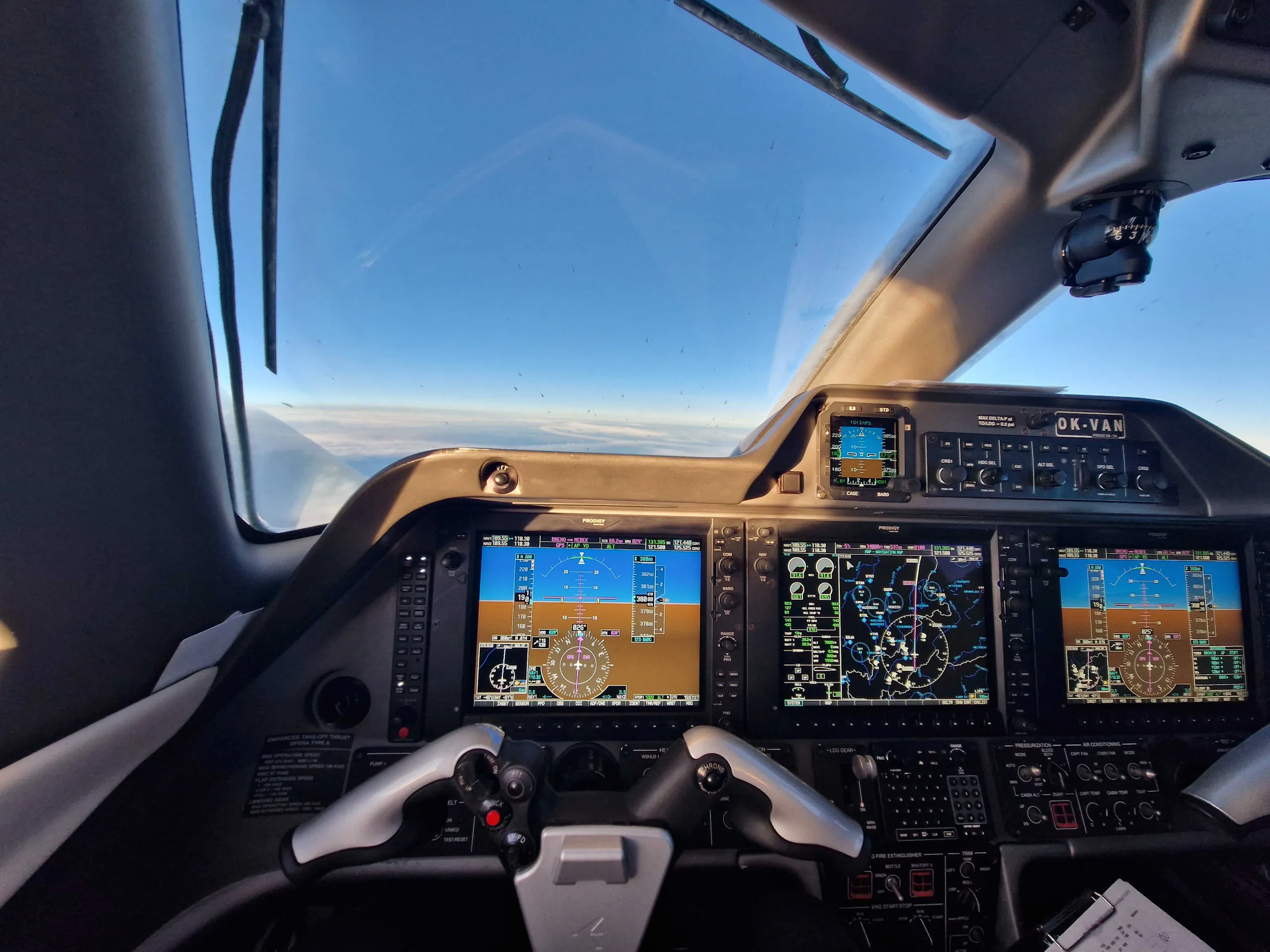 The cockpit of a private jet shows multiple digital displays, control yokes, and the sky visible through the windshield.