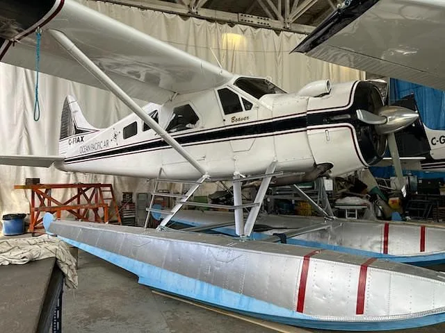 A white De Havilland Beaver seaplane with "C-FIAX" and "OCEAN PACIFIC AIR" markings is seen from the front-left in a hangar.