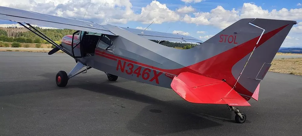 A rear-side view of the gray and red N346X aircraft with "STOL" on its tail, parked on an asphalt apron.