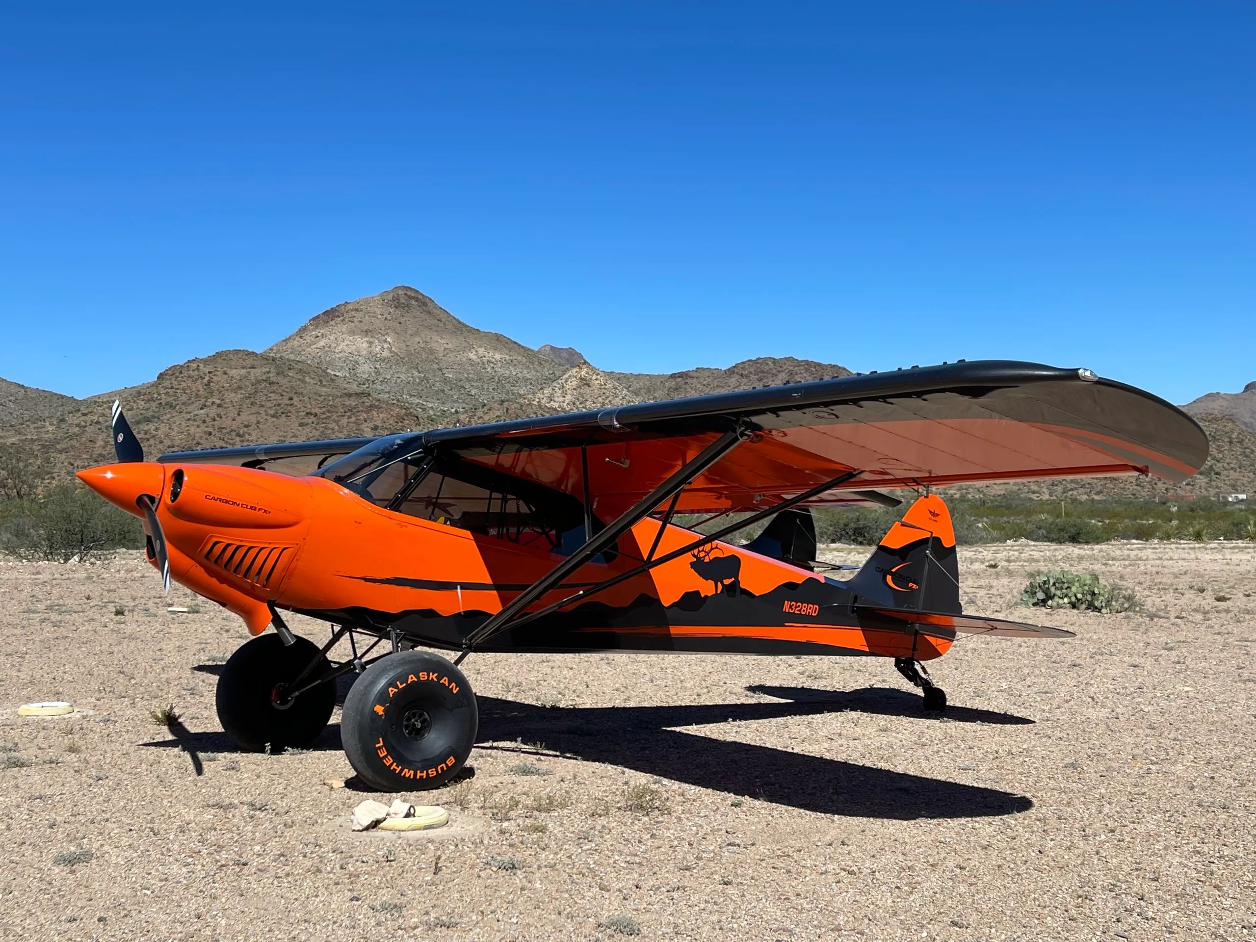 An orange and black Carbon Cub FX-3 aircraft with an elk silhouette on the tail, parked in a desert landscape with mountains.