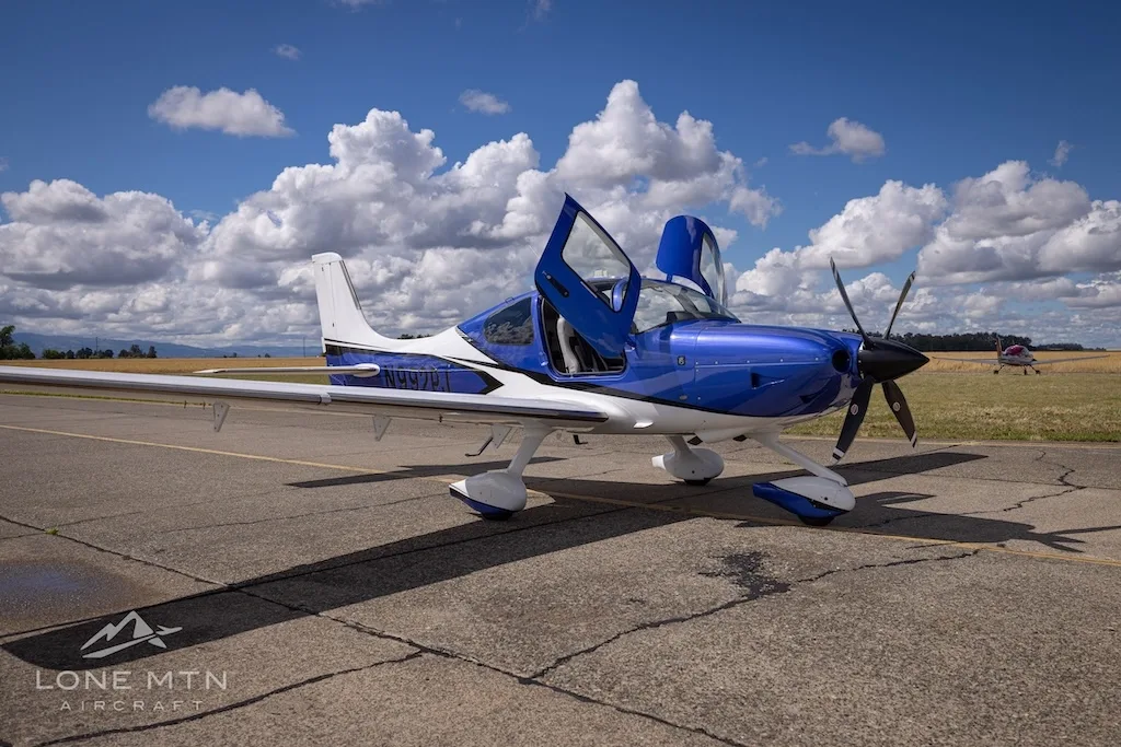 A blue and white Cirrus SR22 aircraft with its doors open, parked on an airfield under a blue sky.