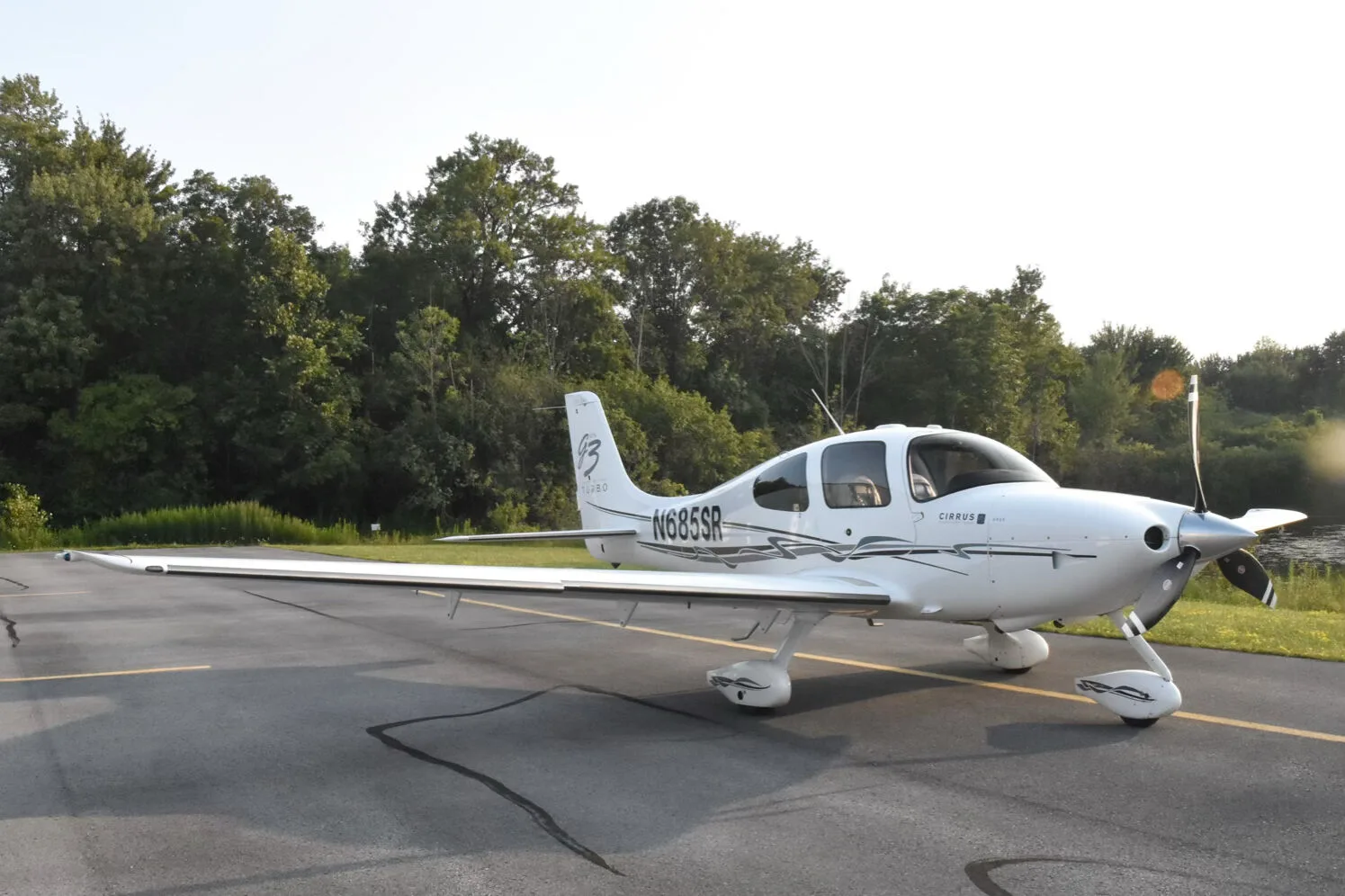 A side view of the white Cirrus SR22T aircraft (N685SR) on an airfield with a line of trees in the distance.
