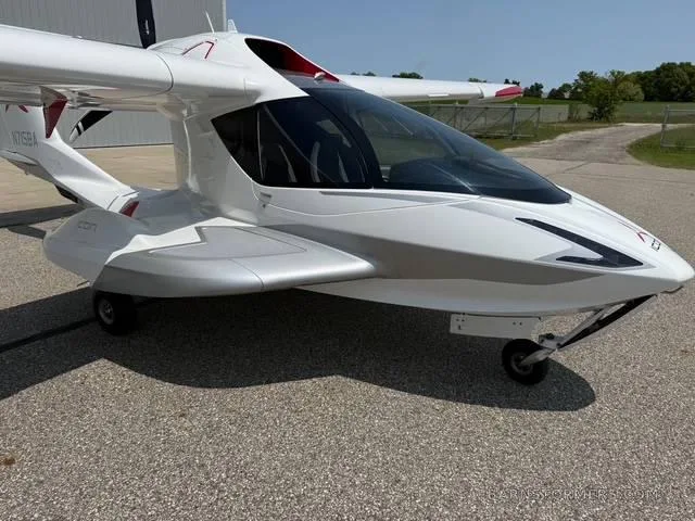 A white and silver ICON A5 amphibious light sport aircraft is parked on an asphalt surface next to a hangar.