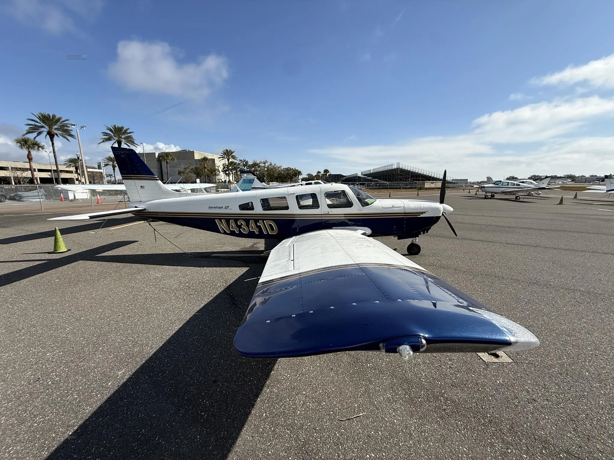 A Piper Saratoga SP (N4341D) in blue and white paint scheme parked on an airfield with palm trees in the background.