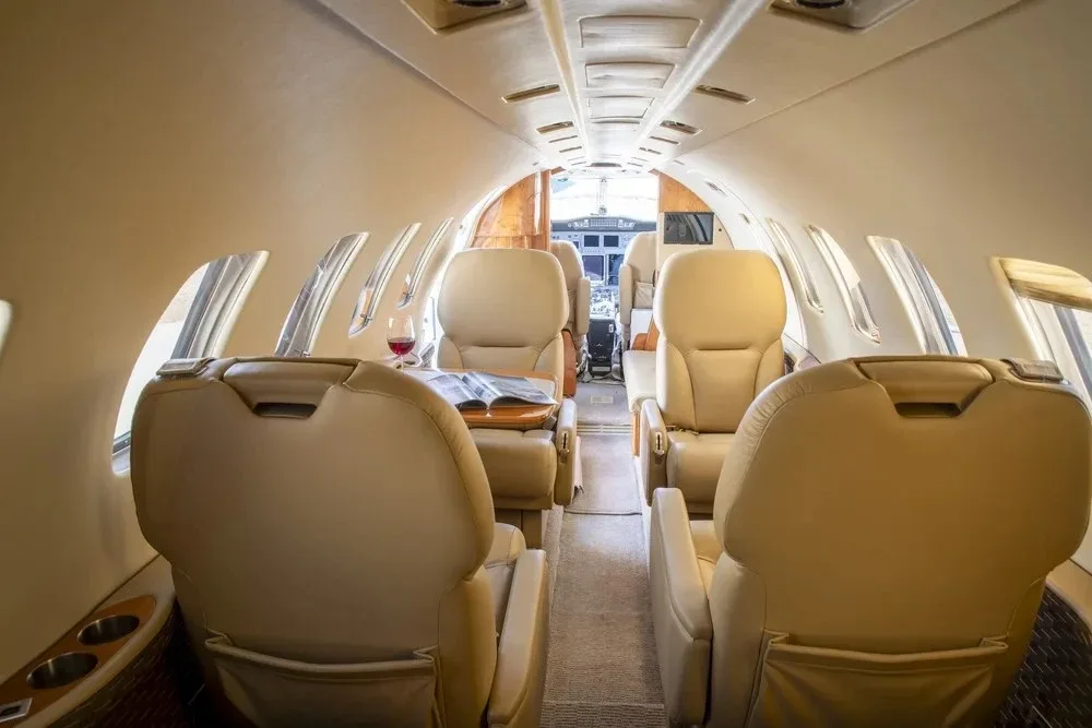 The interior cabin of a private jet featuring light tan leather seats, a small table with a magazine and a glass of red wine, looking towards the cockpit.