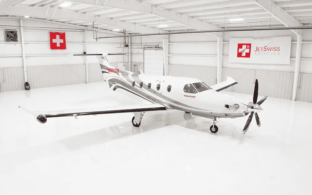 A white Pilatus PC-12 aircraft with gray and red accents is parked in a brightly lit hangar.