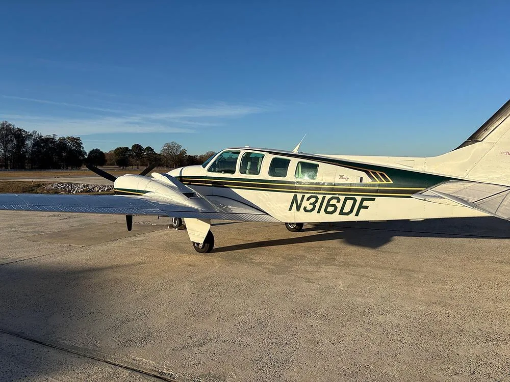 A white and green Beechcraft Baron N316DF parked on an airfield with trees in the background.