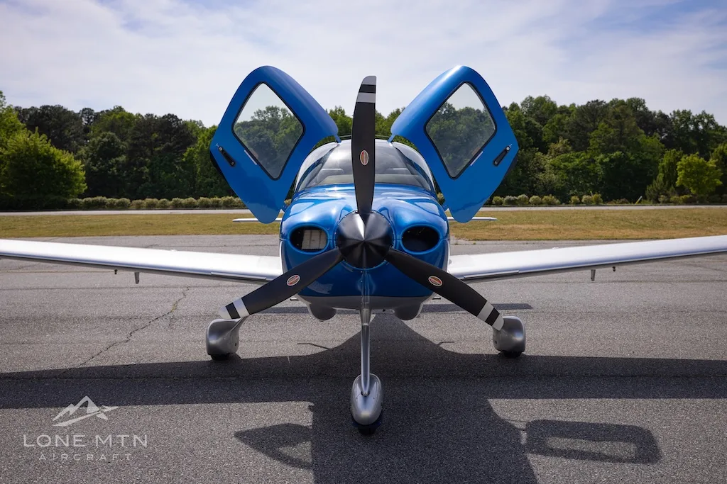 The front view of a blue and silver single-engine aircraft with its two gull-wing doors open.