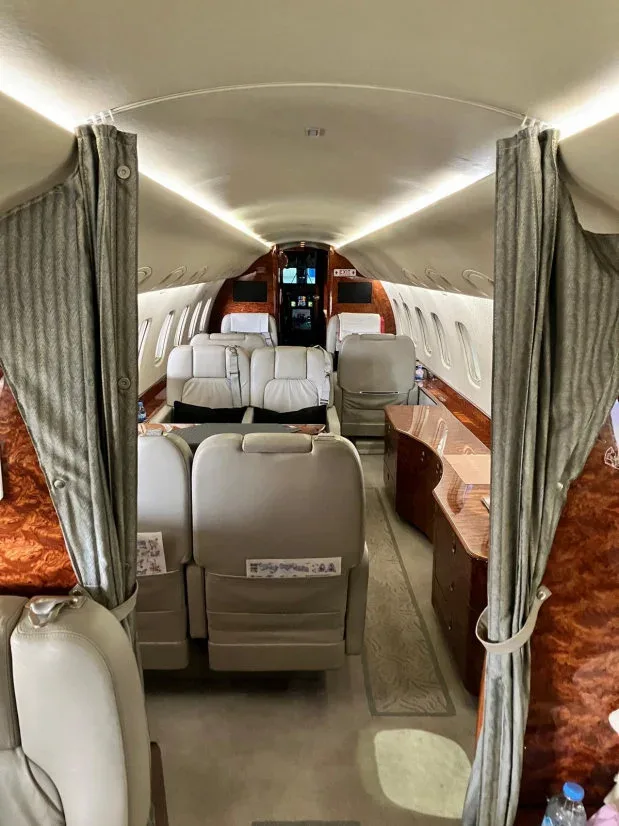 The interior cabin of a private jet is viewed from the rear, showing light grey seats, wood paneling, and open privacy curtains.