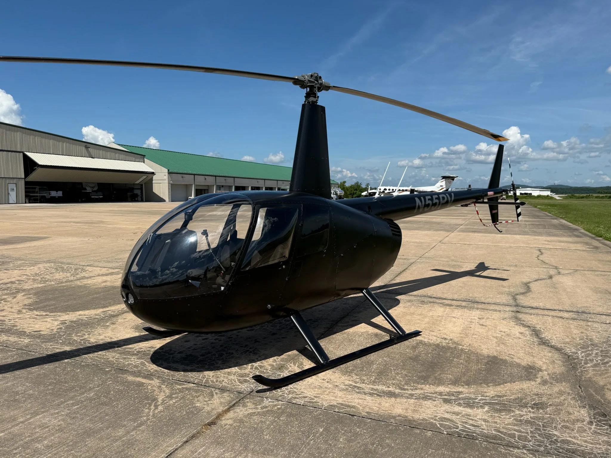 A black Robinson R44 helicopter with registration N55PV is parked on an airport tarmac with hangars in the background under a blue sky.