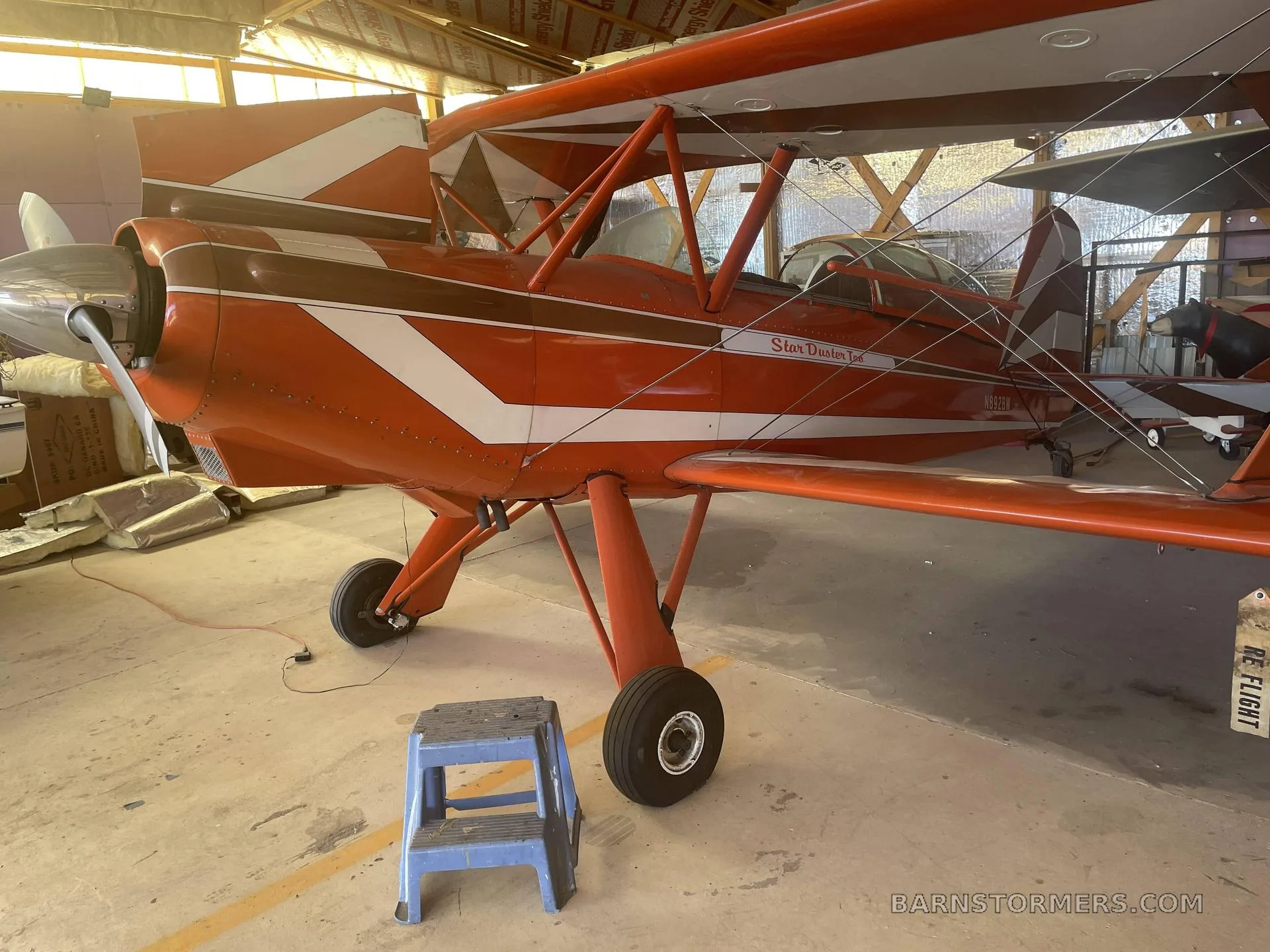 An orange and white Star Duster Too biplane is parked in a hangar with a blue step stool in front.