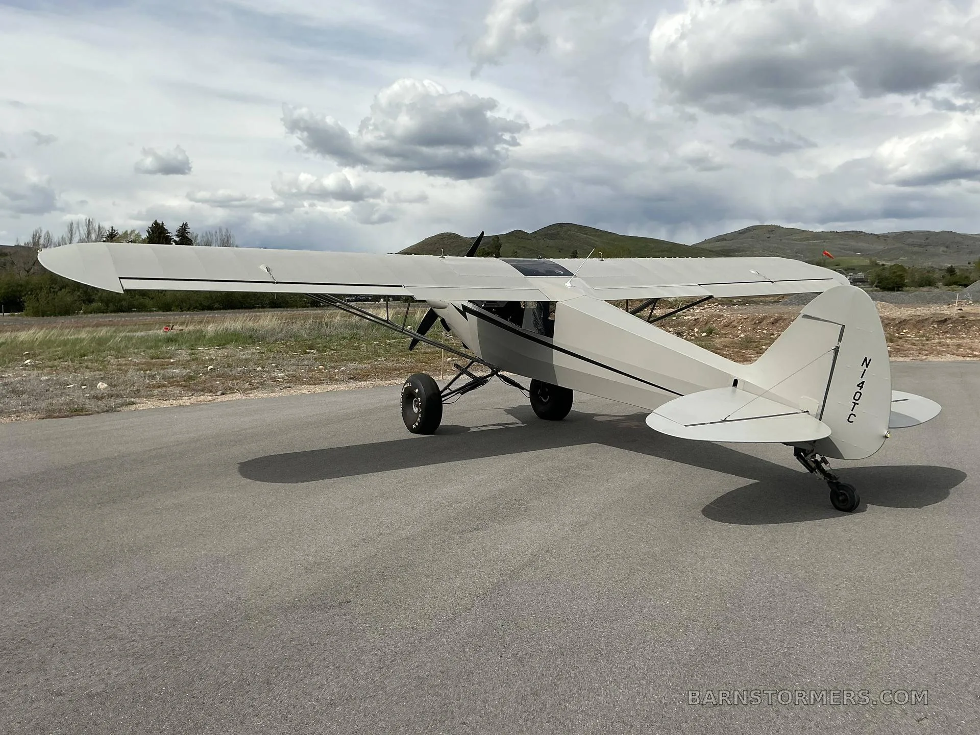A light grey high-wing taildragger aircraft with black stripes and large tundra tires, viewed from the rear-right, parked on asphalt with hills in the background under a partly cloudy sky.